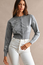 Woman wearing a gray embroidered cardigan with floral and animal designs paired with white high-waisted pants.