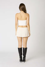 Woman modeling a beige mini skort paired with a white tube top and black knee-high boots from the back view