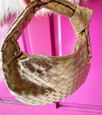Gold woven vegan leather bag with zip closure held against a bright pink background, stylish women's accessory.