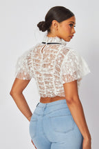 Woman wearing a sheer white lace cropped top with rosette details and high neckline paired with light blue jeans.