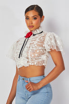 Woman wearing a sheer white lace crop top with puff sleeves and a red rosette detail paired with light blue jeans