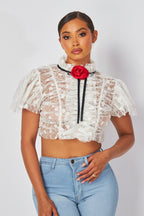 Woman wearing sheer white lace crop top with puffed sleeves and red rosette detail paired with light blue jeans