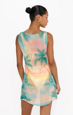 Woman wearing a sleeveless tropical print dress with palm trees and sunset in pastel colors from the back view