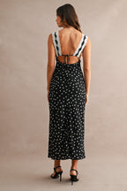 Woman wearing black polkadot maxi dress with lace straps and open back, styled with black heels, standing against beige wall.