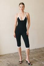 Woman wearing black capri jumpsuit with white lace trim and deep V neckline in a minimal indoor setting