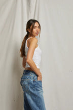 Woman wearing a fitted white tank top and relaxed blue denim jeans standing against a neutral background