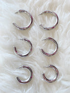 Three pairs of chunky silver hoop earrings in varying sizes displayed on soft white fabric background