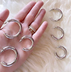 Hand holding various sizes of chunky silver hoop earrings displayed on soft white fabric background