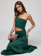Woman wearing a ruched, one-shoulder green maxi dress seated on the floor holding a brown clutch bag