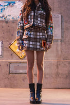 Woman wearing a fitted plaid shacket with colorful patches paired with a matching skirt and black ankle boots.