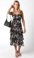 Woman wearing a strapless tiered floral midi dress holding a black handbag and wearing black heeled sandals.