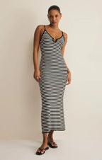 Woman wearing a black and white striped midi dress with thin straps in a minimalist setting standing barefoot.