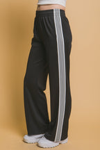 Women's black track pants with white side stripes, elastic waistband, and relaxed fit paired with white sneakers.
