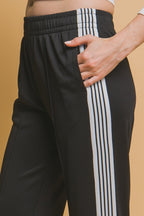 Close-up of black relaxed track pants with white side stripes and a hand in the pocket, styled for women's clothing.