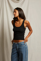 Woman wearing a fitted black tank top with wide straps paired with blue denim jeans against a neutral background