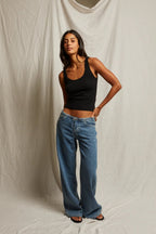 Woman wearing a fitted black tank top and loose blue jeans standing against a neutral fabric backdrop