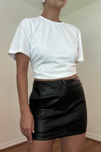 Woman wearing a black faux leather mini skirt paired with a plain white cropped t-shirt in a simple setting