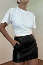 Woman wearing a black faux leather mini skirt with a white fitted cropped t-shirt, hand in pocket, fashion pose.