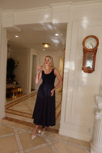Woman wearing a sleeveless black midi dress with a fitted bodice and full skirt holding a glass in an elegant room