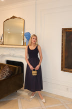 Woman wearing a sleeveless midi dress with a fitted bodice and flowing skirt holding a small handbag in a stylish room.