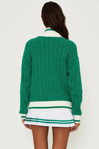 Woman wearing oversized green cable knit sweater with ribbed cuffs and hem, paired with white pleated skirt