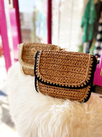 Brown woven crossbody bag with black vegan leather trim displayed on a white fur surface in a boutique setting