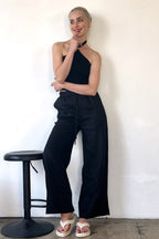 Woman wearing a black ribbed halter top paired with loose black pants and white sandals against white wall.
