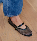 Woman wearing black fishnet Mary Jane ballet flat with buckle and blue jeans on wooden floor