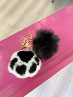 Two faux fur poof keychains with gold clasps, one black and one black-and-white spotted, on a pink surface
