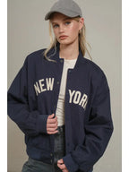 Woman wearing oversized navy varsity jacket with bold New York lettering, white top, gray cap, styled casual women's clothing