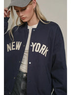Woman wearing a navy oversized varsity jacket with New York lettering and ribbed cuffs, styled with casual clothing