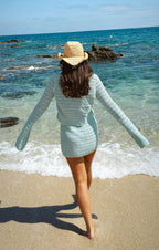 Woman wearing a light blue knit skirt walking barefoot on the beach with a straw hat and long sleeves top
