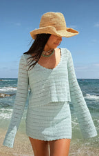 Woman wearing light blue crochet top and skirt with wide-brimmed hat on beach by ocean waves