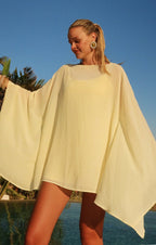 Woman wearing a light yellow flowing top with wide sleeves standing outdoors near a pool under clear blue sky