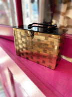 Vintage brass woven bag with black handle and latch closure displayed on pink surface for women's fashion accessory.