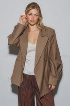 Woman wearing a brown oversized blazer with wide lapels over a white top and textured brown pants in a studio setting