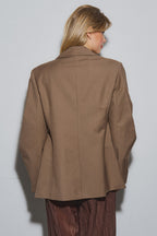 Woman wearing a brown oversized blazer from the back, styled with loose brown pants, against a plain gray wall.