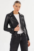 Woman wearing a fitted black leather moto jacket with a diagonal zipper and snap collar over a white top.