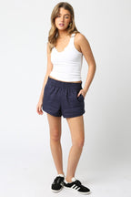 Woman wearing a white sleeveless tank top paired with navy casual shorts and black sneakers in a studio setting.