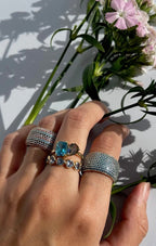 Hand wearing layered gold rings with blue and green gemstones holding pink flowers against a white background.