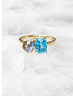 14K gold ring with a vibrant blue square gemstone and a gray-blue oval gemstone on a white background