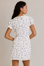 Woman wearing a white mini dress with blue floral print and puff sleeves, shown from the back, modeling women's clothing.