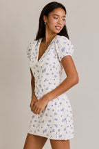 Woman wearing a white mini dress with small blue floral print and puff sleeves, posing sideways against plain background