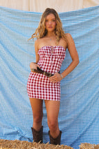 Woman wearing red and white gingham strapless mini dress with brown belt and boots in front of blue gingham backdrop