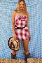 Woman wearing a red and white gingham strapless mini dress with a black belt, holding a cowboy hat and brown boots