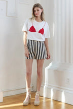 Woman wearing white tee with red bikini print and black and white striped high-waist shorts in bright room