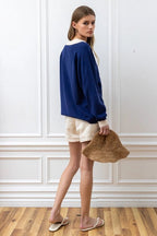 Woman wearing a navy knit pullover with cream cuffs and collar, paired with beige shorts and holding a straw hat