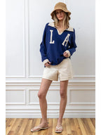 Woman wearing a navy knit pullover with white LA letters, beige collar, cuffs, shorts, and a woven bucket hat