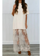 Woman wearing a cream shirt dress with sheer lace maxi skirt overlay and brown pointed heels in a minimalist room