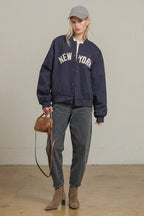 Woman wearing navy bomber jacket with "New York" text, gray jeans, gray cap, and brown ankle boots holding a small bag